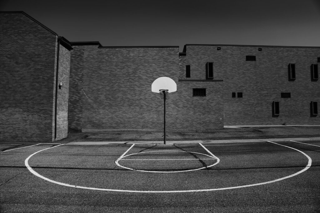 Basketballplatz in Schwarz-Weiß. Im Vordergrund sind die Linien des Spielfels, die einen Halbkreis und das Freiwurfviereck markieren. In der Mitte steht ein Basketballkorb mit einem Netz.